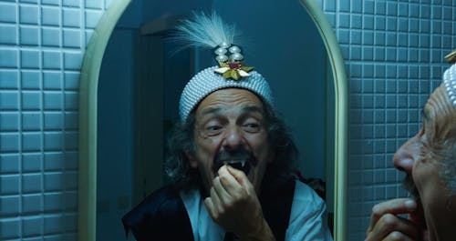 In a blue-lit tiled room, a man with gray curly hair and a mustache touches his long incisor fang while looking in the mirror. He wears a jeweled hat.