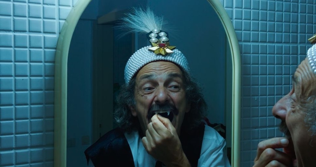 In a blue-lit tiled room, a man with gray curly hair and a mustache touches his long incisor fang while looking in the mirror. He wears a jeweled hat.