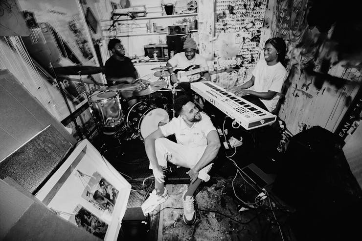 Grayscale image of four musicians playing in a small room with tons of music gear