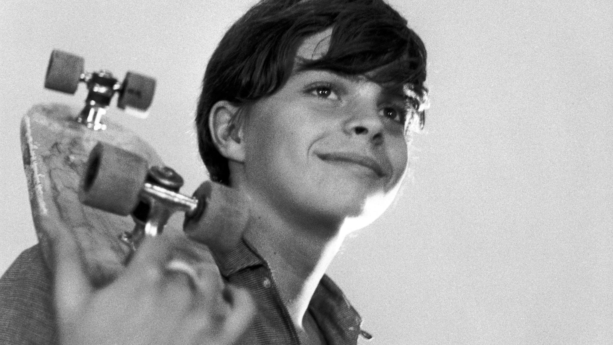 Black and white image of a boy smiling holding a skateboard over his shoulder.