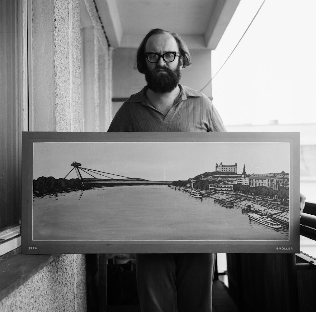A man with a beard and glasses stands on a balcony of an aprtment block holding a painting of a city on a river while looking at the viewer.