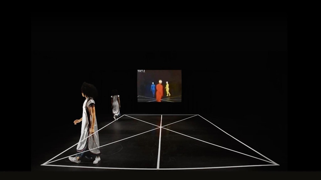 A dancer in white walks along the edge of a dance floor, her body doubled in the space. A projection along the back wall shows three figures in primary colors.