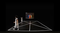 A dancer in white walks along the edge of a dance floor, her body doubled in the space. A projection along the back wall shows three figures in primary colors.