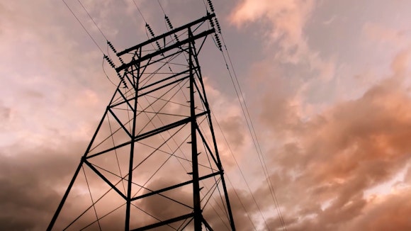 A high power line tower stands in front of a clowdy sky.