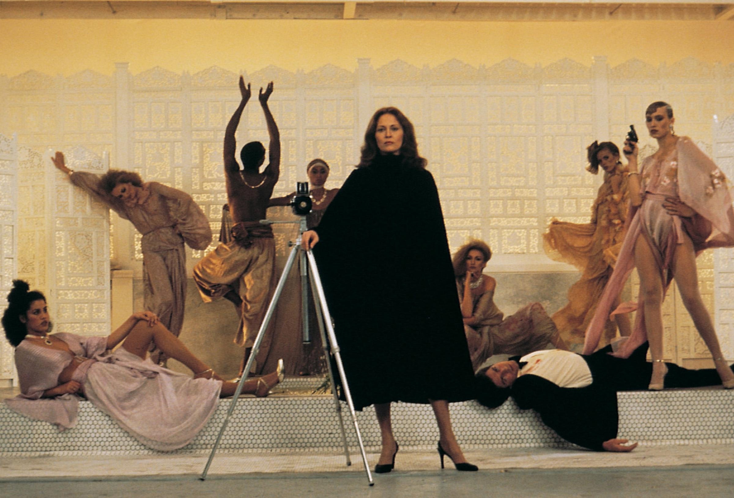 A photographer in black stands with a tripod on a set among models in flowy golds and pinks. A man lies dead on the floor.