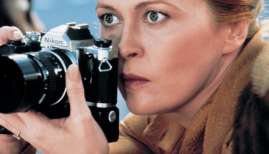 A close shot of a photographer in brown and fur clothing looking intensely out from behind her camera.