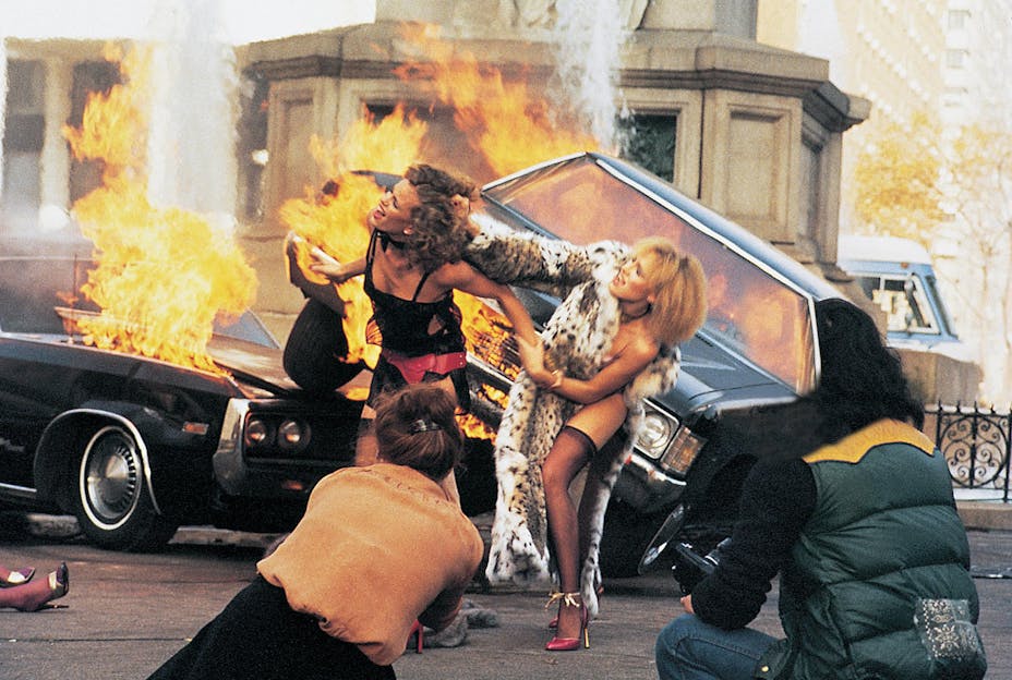 Two models fight physically with each other in front of burning cars while photographers capture the scene.