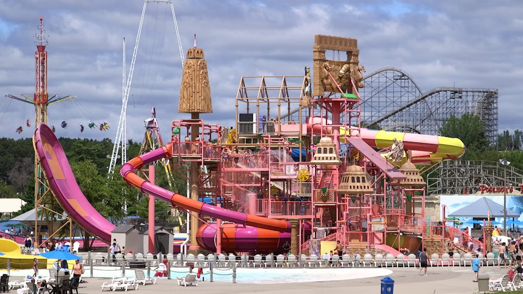 A photo of an entire outdoor waterpark, in bright colored in pink, yellow and orange. There are roller coasters and swing rides in the background.