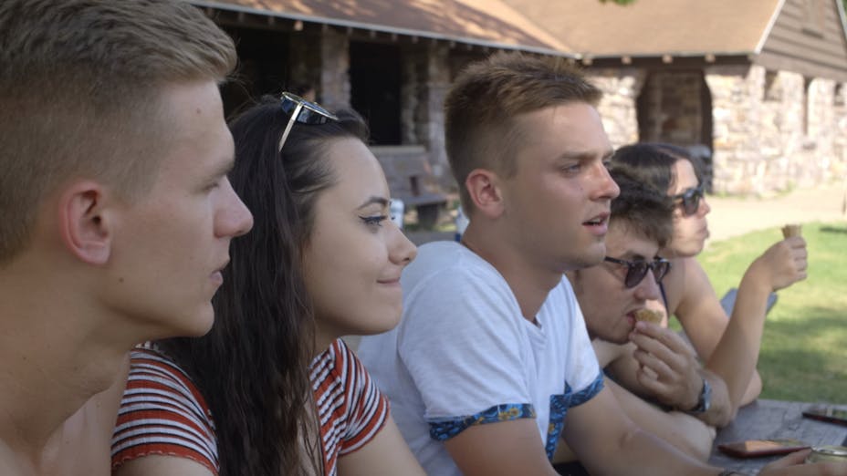 Five young adults with light skin tones sit at a picnic table in front of a stone park pavilion. They are talking and eating ice cream.