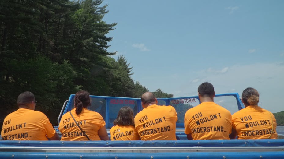 Six people in a blue boat wear the same yellow tshirt that reads “YOU WOULDN’T UNDERSTAND...” on the back. The water is surrounded by trees.