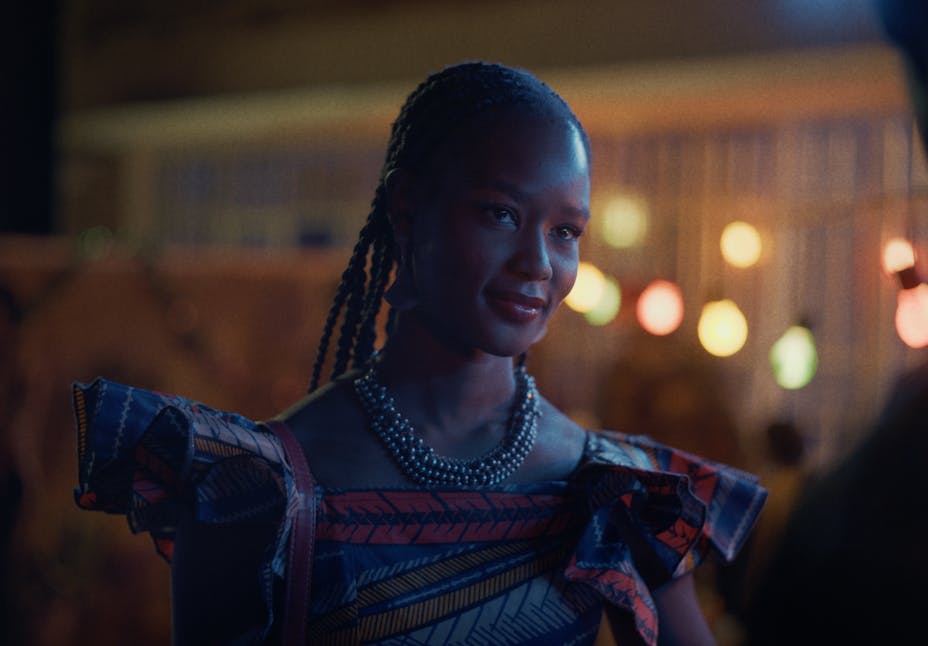 An adult with braids and dark skin stands outside and looks away at the viewer.