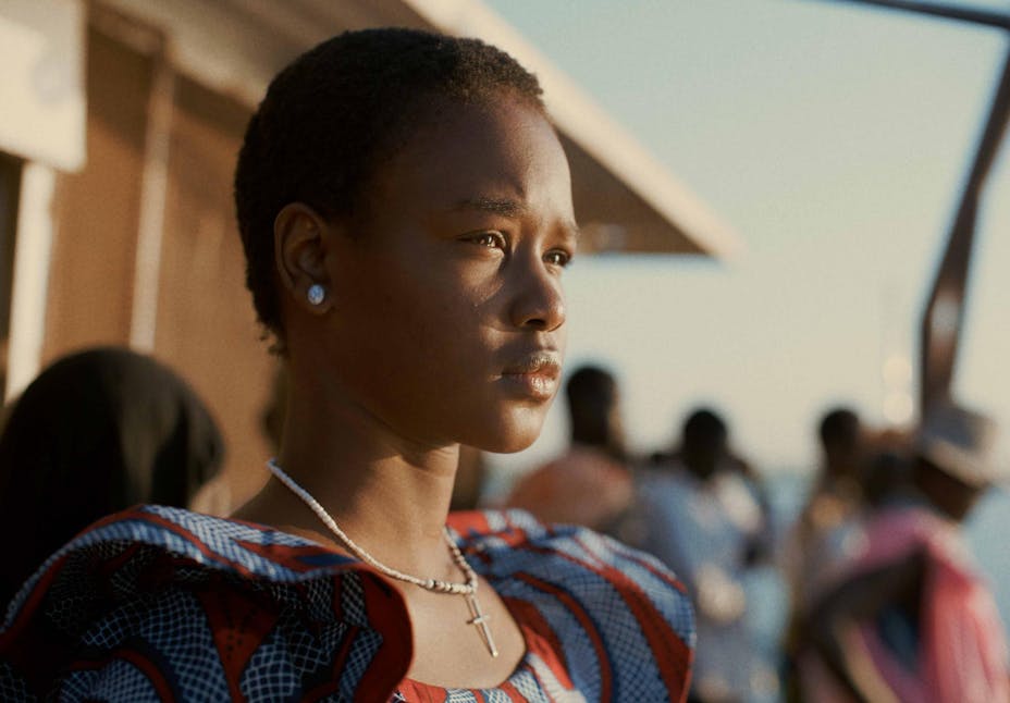 An adult with braids and dark skin stands outside and looks away at the viewer.