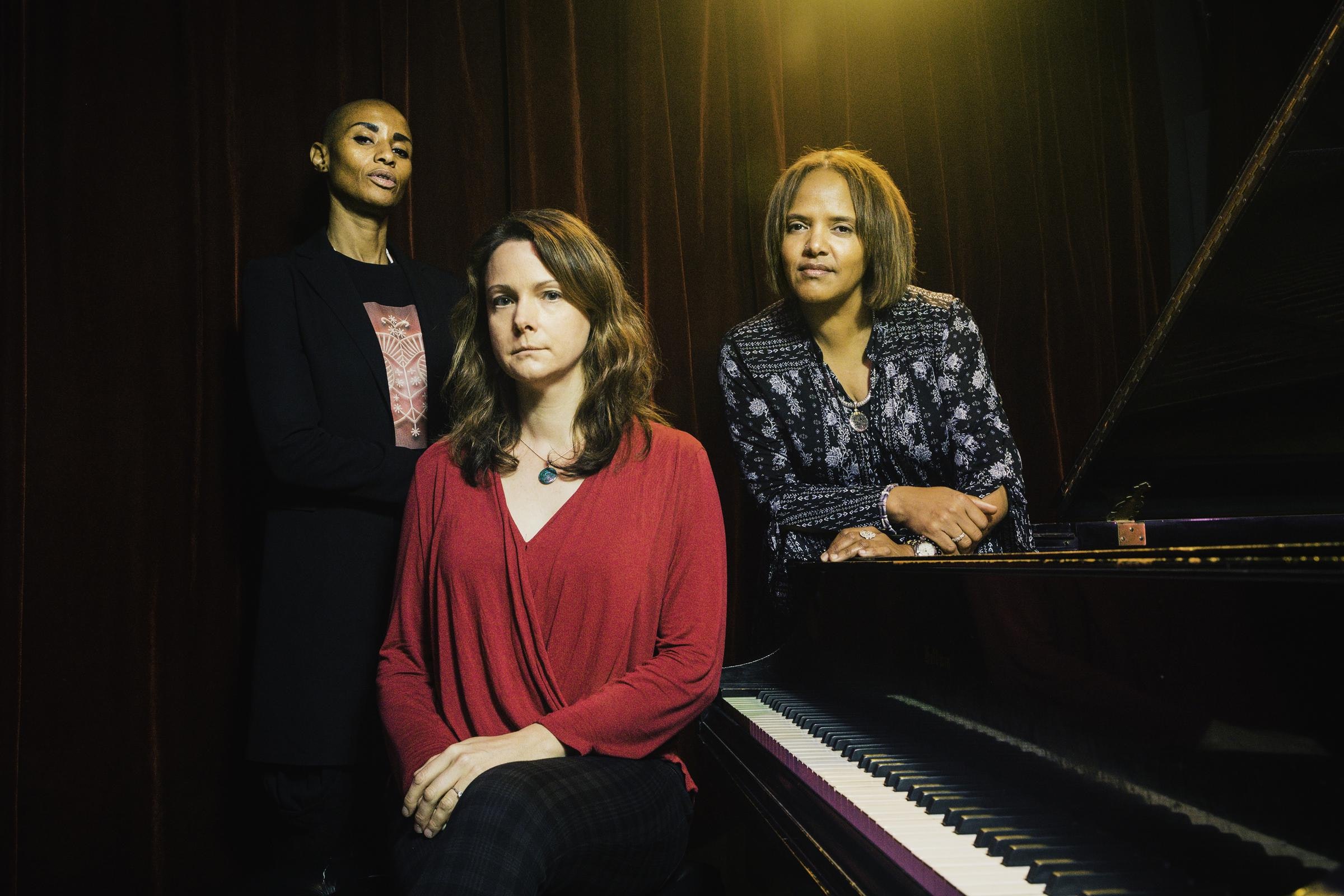 Three women stand around a piano.