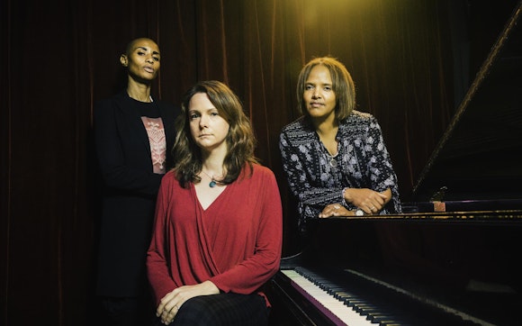 Three women stand around a piano.