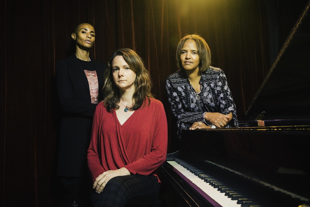 Three women stand around a piano.