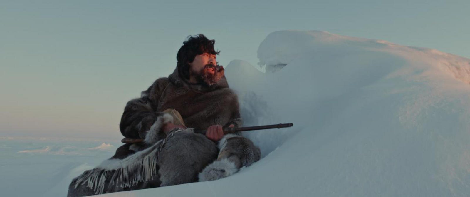 A man with dark hair wearing furs sits against a snow formation and looks out over it holding a shotgun. There is dim sun over the snow covered landscape.