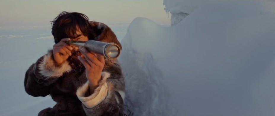 A man with dark hair wearing a fur coat holds a telescope to his eye. He sits next to a snow and ice formation on a totally snow-covered background.