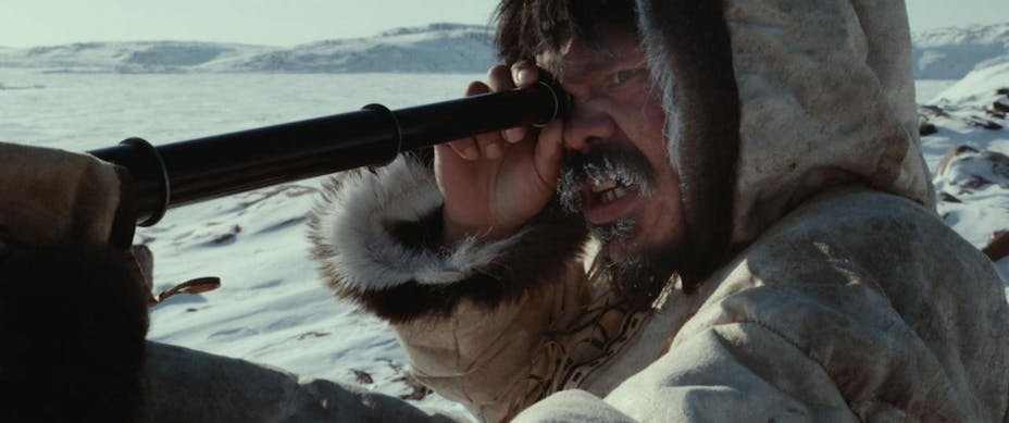 A man in a leather fur-lined cat holds a telescope to his eye in an arctic landscape. He has an intense scowl and ice is forming on his facial hair.