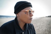 An adult with dark skin looks away from the viewer while wearing sunglasses and a black knitted cap on a beach