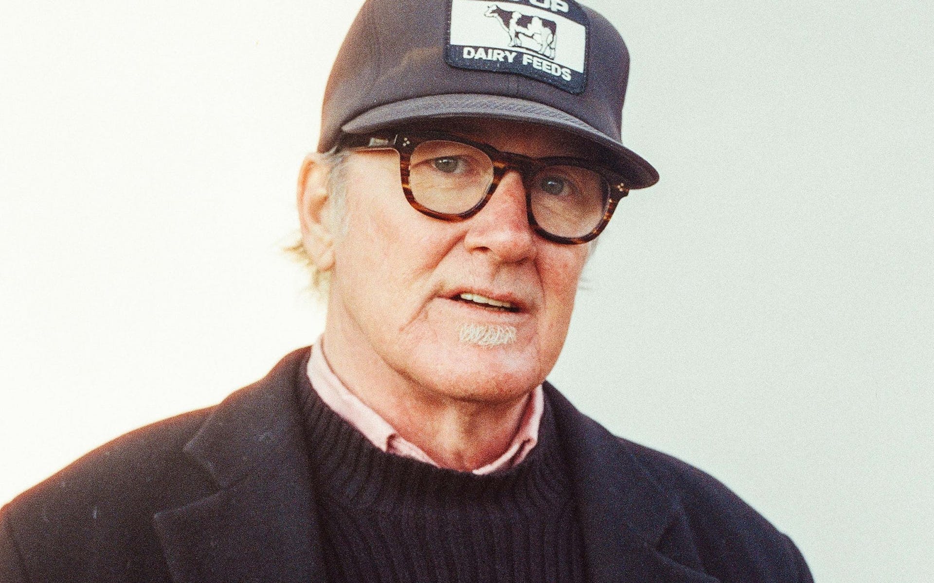 A man in a black baseball cap, cuitcoat, and glasses looks at the viewer.