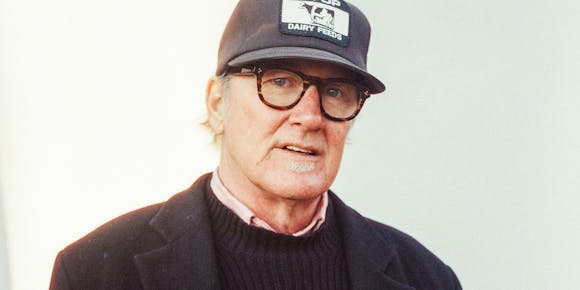 A man in a black baseball cap, cuitcoat, and glasses looks at the viewer.