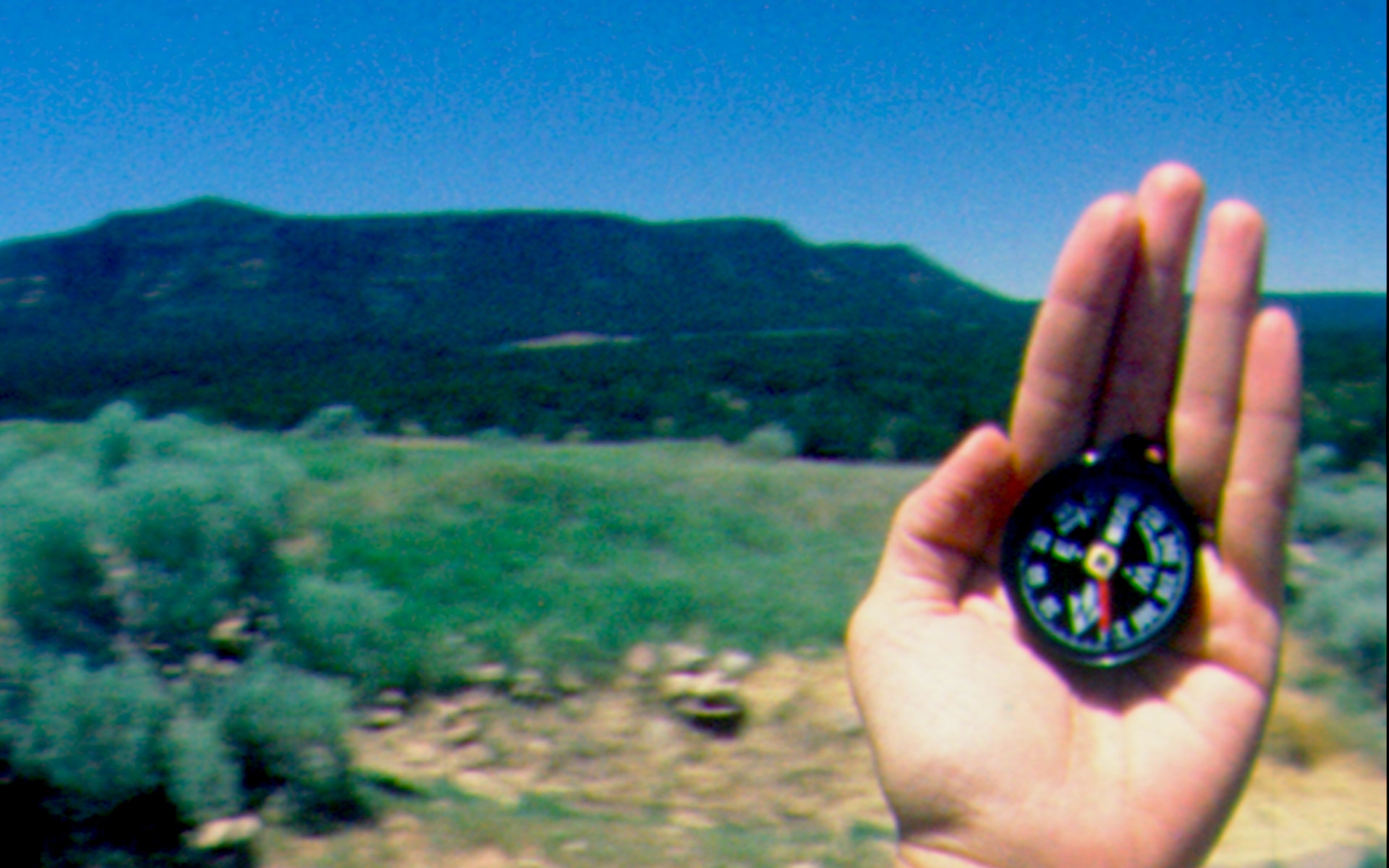 A hand holds up a compass in front of a mountain vista.