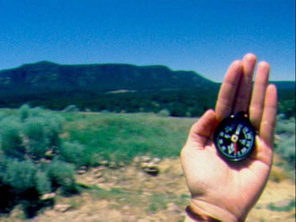 A hand holds up a compass in front of a mountain vista.