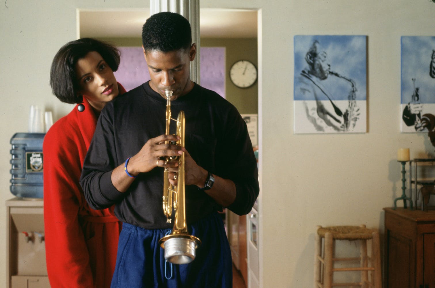 A man looking down plays trumpet while a woman in a red coat behind him rests her chin on his shoulder.