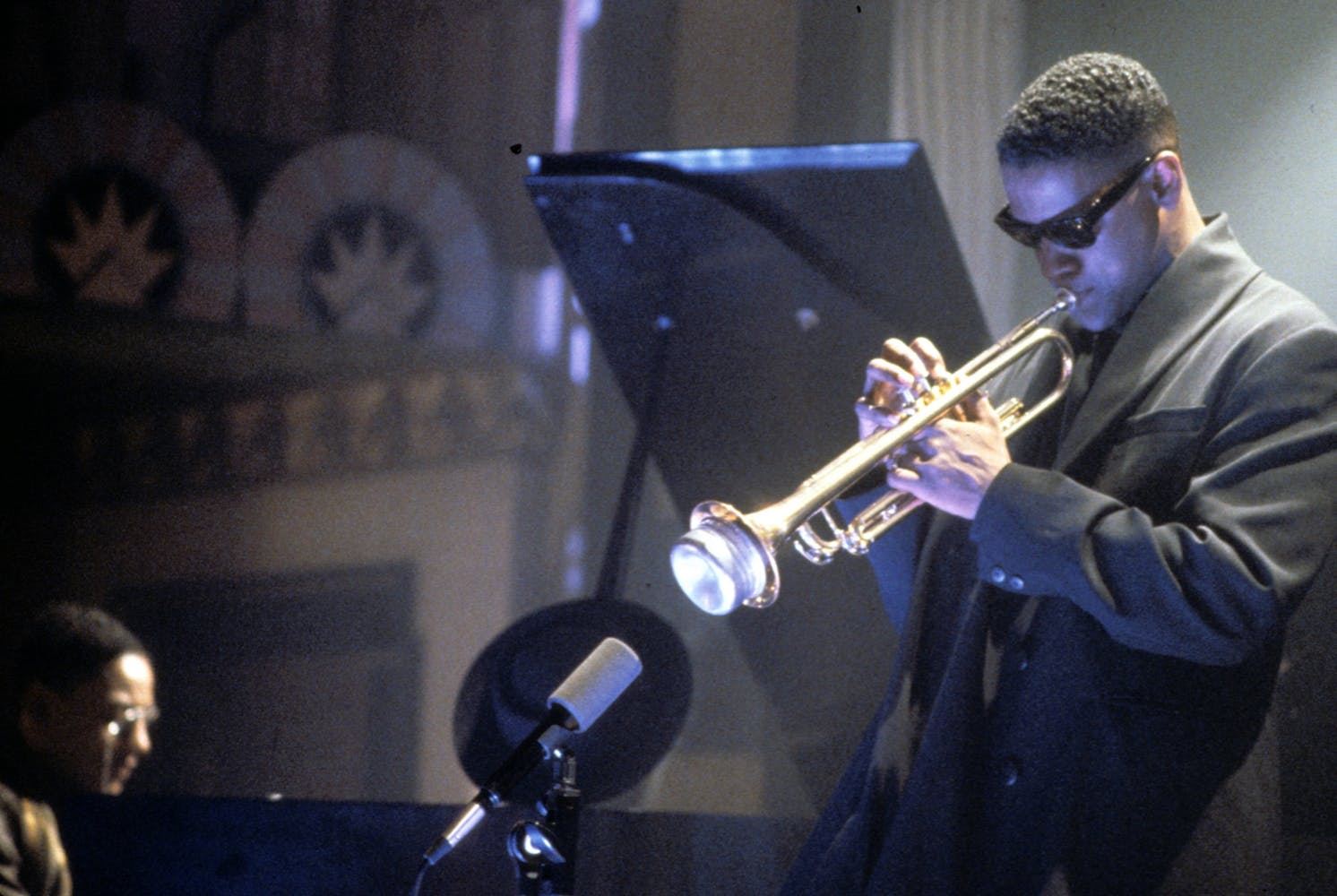 A man in a black coat adn sunglasses plays trumpet on a stage next to a piano player.