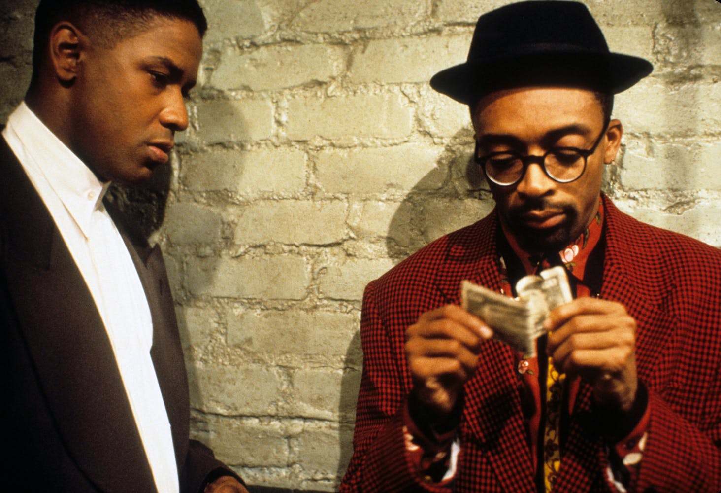 A man stands next to another man in a hat and glasses who is holding money in a hallway.