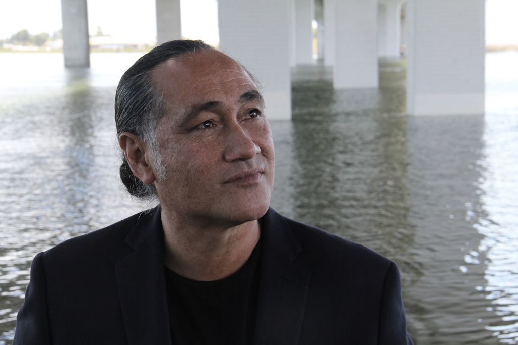 A man wearing a blaze blazer witht heri hair pulled back looks off in 3/4 profile from the viewer with water behind them.