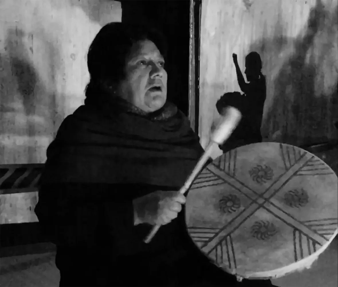 An older woman sings while playing a handheld drum with the shadow of a dancer behind her.
