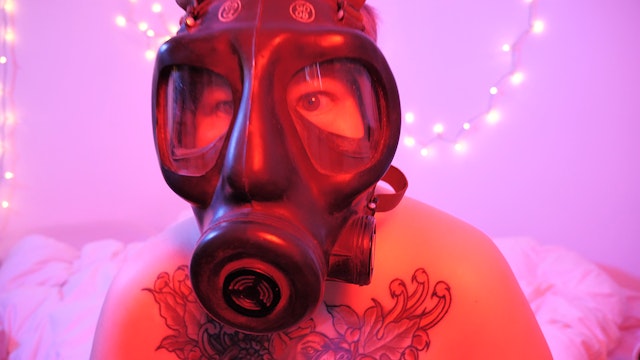A close up of an adult wearing a gas-mask faces the viewer in a red an pink lit room
