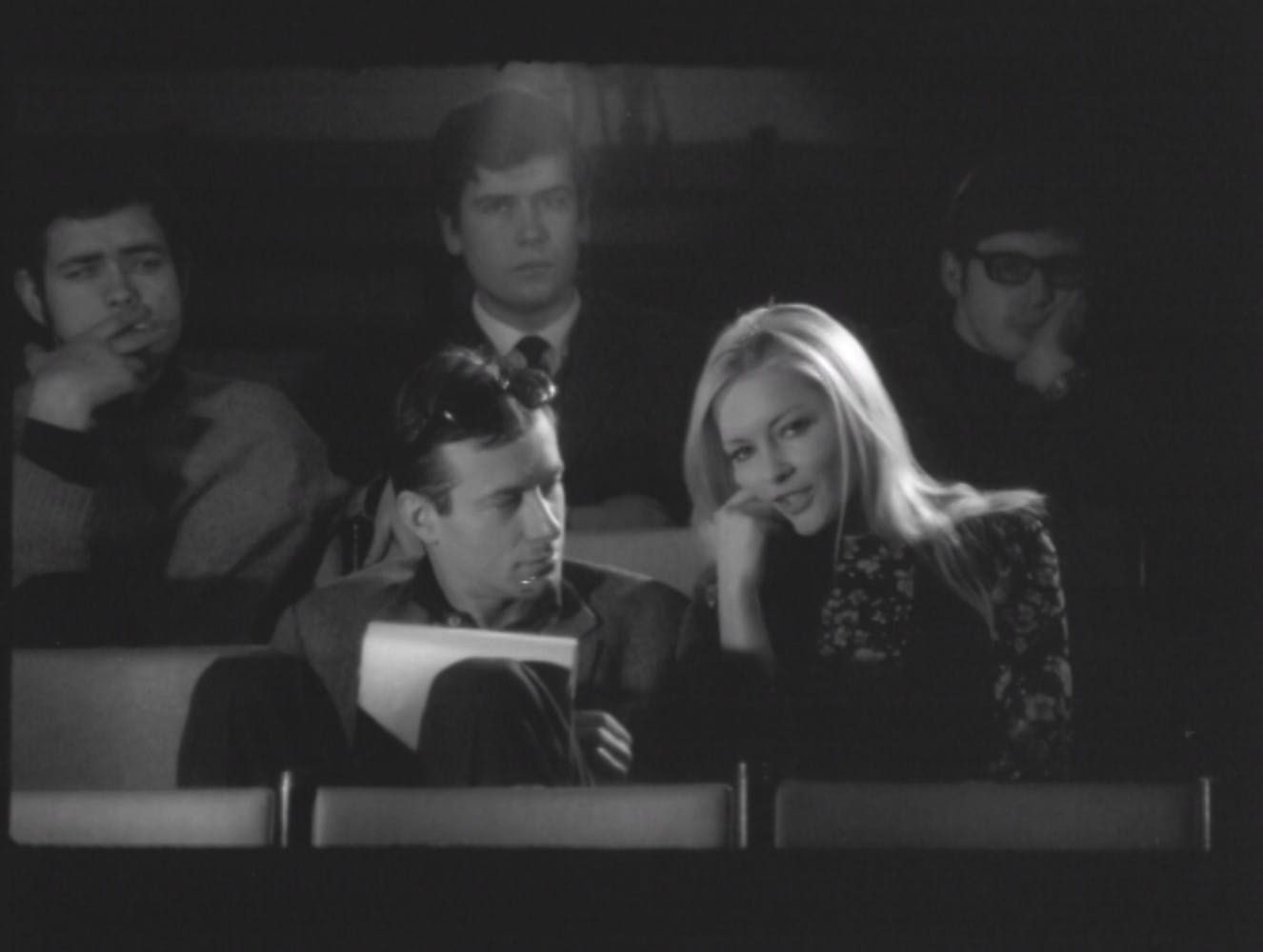 A woman with long light hair and light skin speaks to a man with light skin, dark hair, and dark sunglasses on her head as they sit in a movie theater.