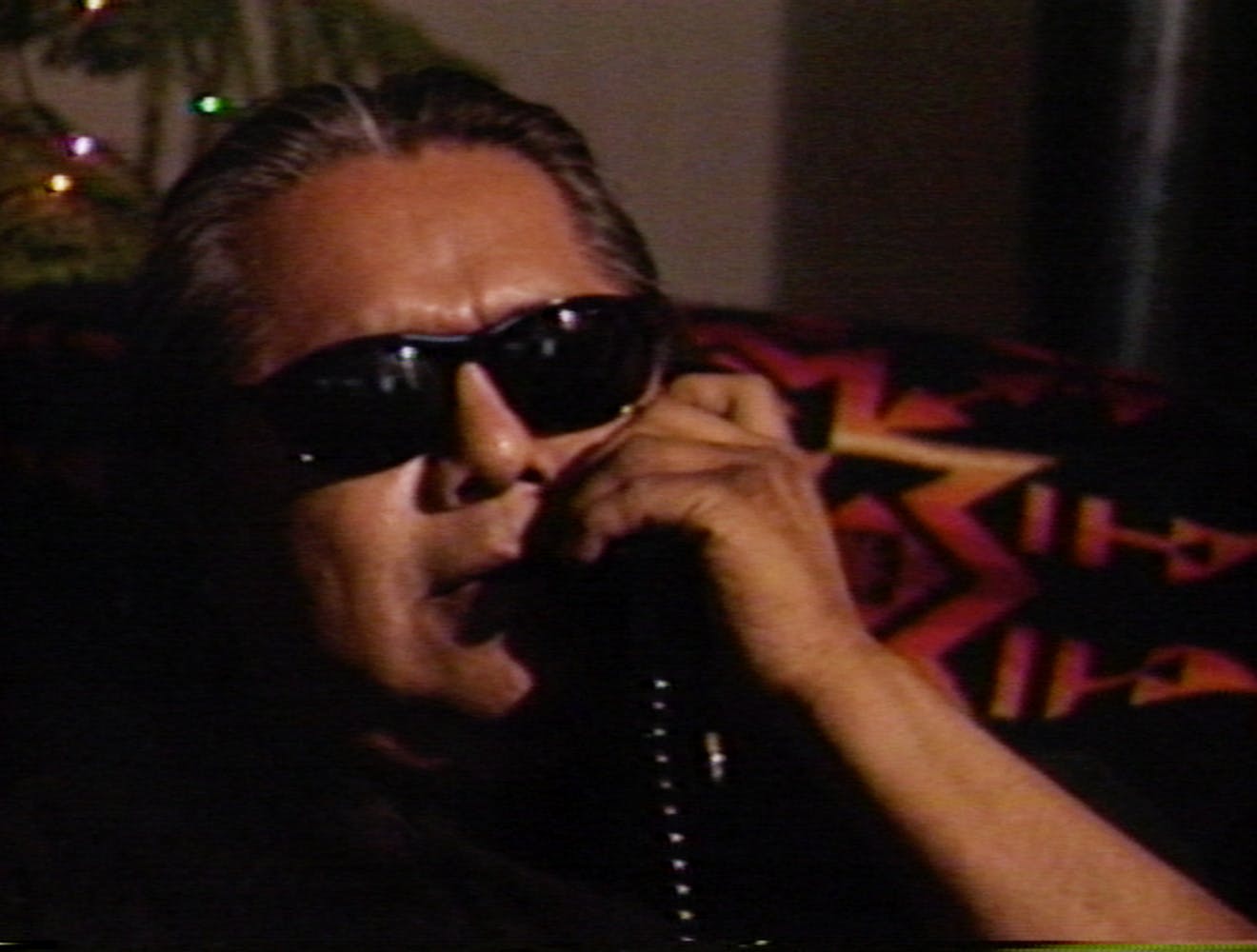 A close image of a man sitting, wearing sunglasses, and holding a corded phone to his ear. Behind him is a patterned blanket.