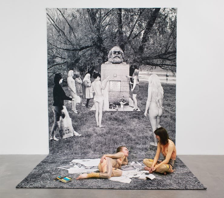 Two women wearing nude jumpsuits sitting on a tapestry that hangs from the wall onto the floor. The tapestry is a black and white image of several women gathered around a gravestone with a sculpture of Karl Marx head on top.