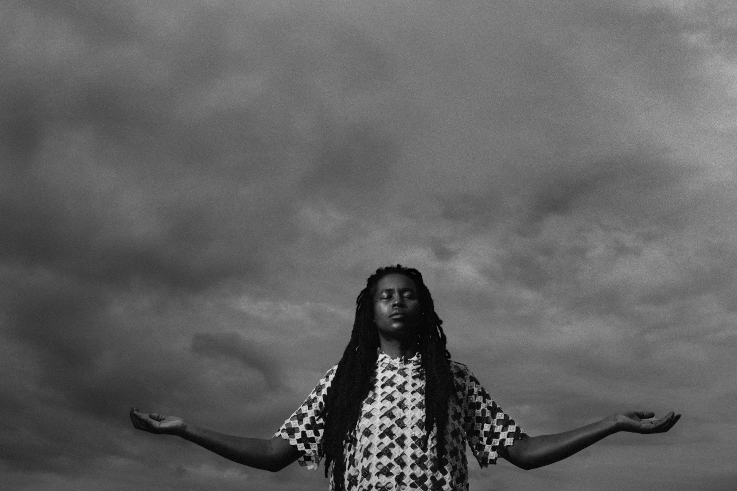 A person with dark skin stands in front of a cloudy sky with their eyes closed and arms outstretched.