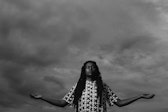 A person with dark skin stands in front of a cloudy sky with their eyes closed and arms outstretched.
