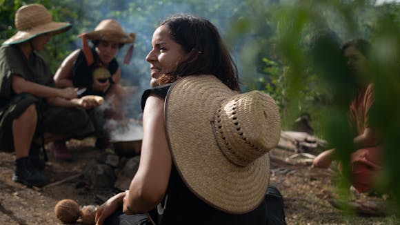 A person with a large straw hat on their back sits in a circle of adults around a fire in nature.