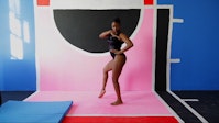 A gymnast poses in a room with graphic shapes on the floor and wall, in black, pink, blue, and red with white lines.