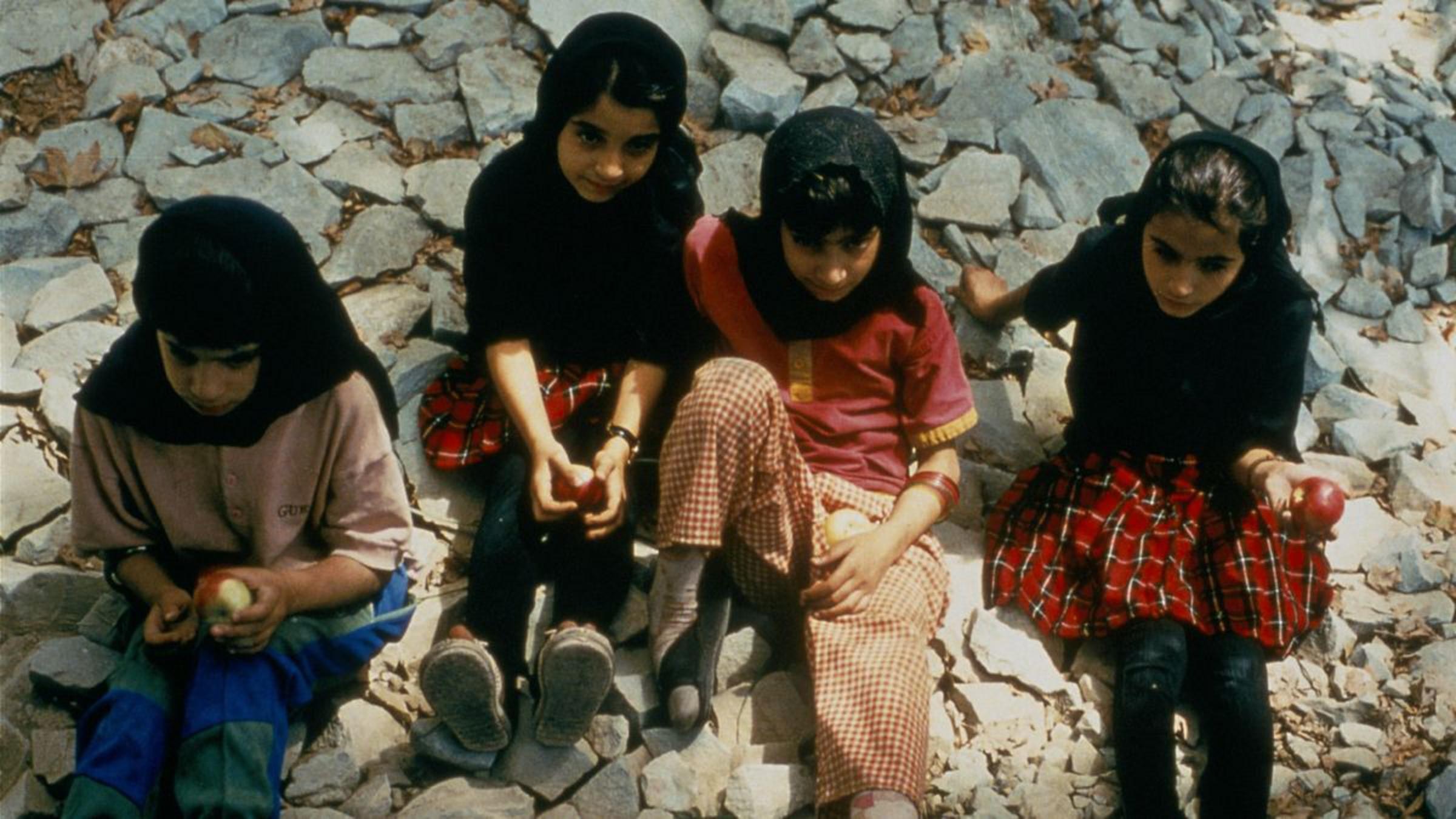 An image looking down toward four kids with dark hair, medium skin tone, and headscarves, sitting on rocks and holding apples.