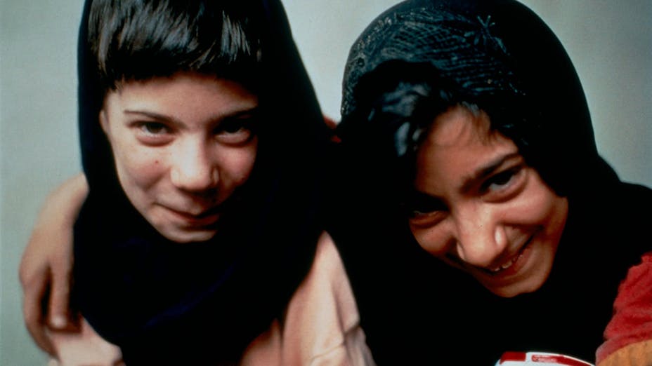A close up of two children with their arms around each other smiling up at the camera. They have dark hair and wear headscarves.