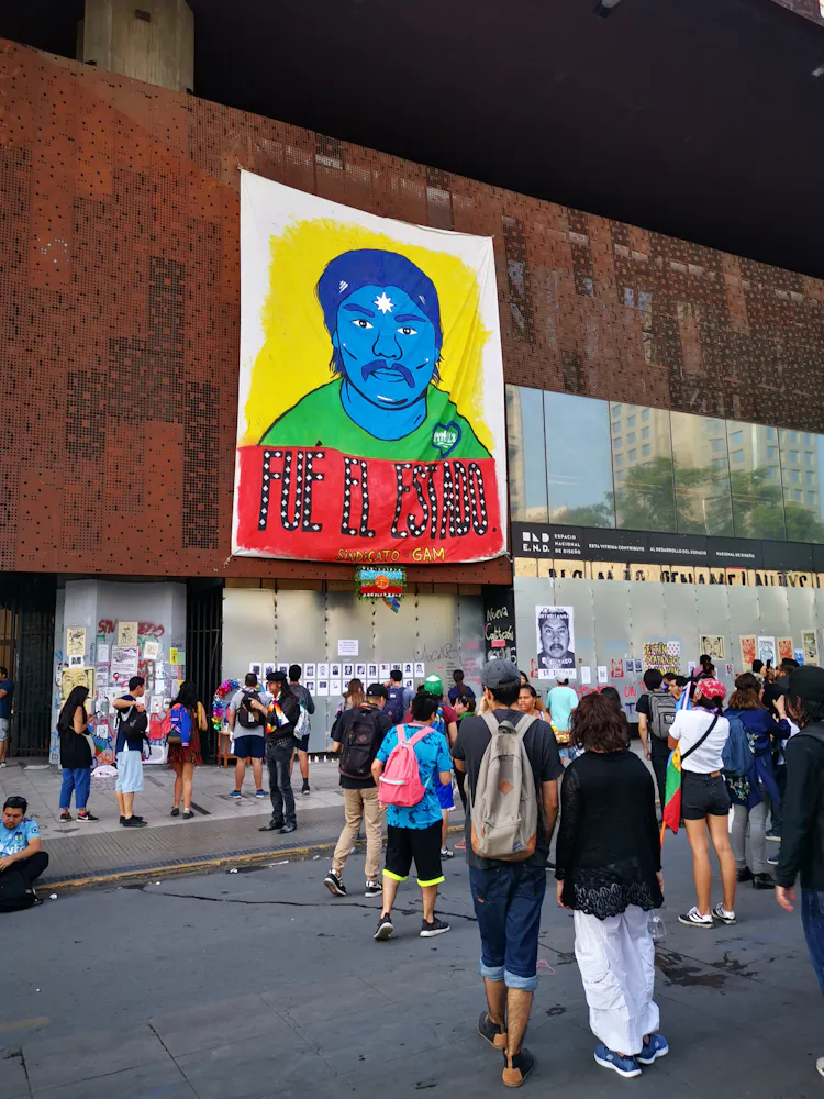 A group of people protest outside of of a community center that has had a large painting of a man's face hung on it.