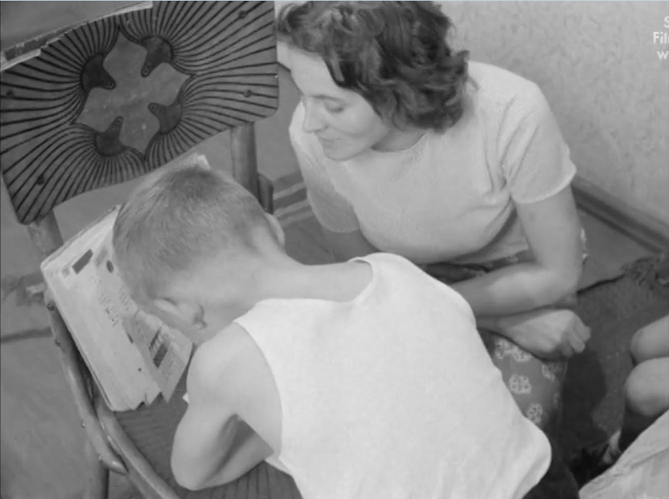 A woman with light skin sits next to a boy with light skin as he does his homework on a chair.