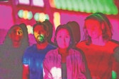 Four performers stand facing the camera in pink light.