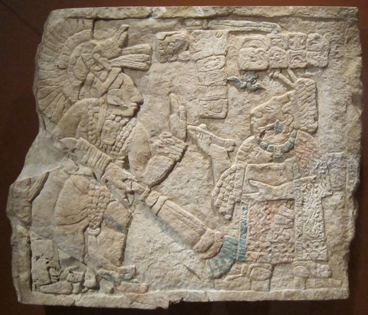 This image shows a square stone Mayan ruin affixed to a dark wall in a museum setting. The tone of the ruin is tan with areas of faded blue and orange color. The ruin depicts hieroglyphic writing and two Mayan figures, one sitting, one standing, interacting formally.