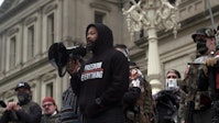 An adult with dark skin wears a black hoodie stands in front of a captial building with a megaphone.