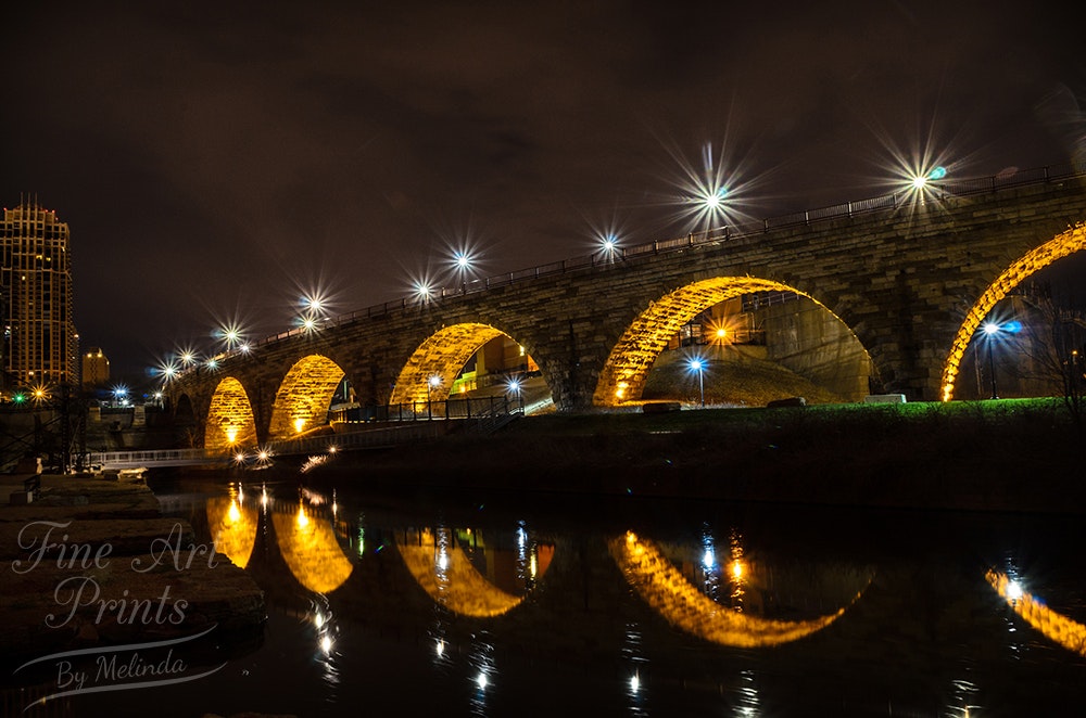 A stone arch bridge at night. There are bright street lights shining on top of the bridge and within the arches below, which reflect off the water