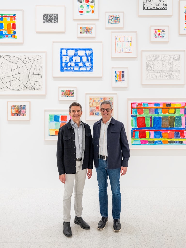 Two adutls with blue jackets and button up shrits smile facing the viewer in front of colorful paintings in a gallery space.