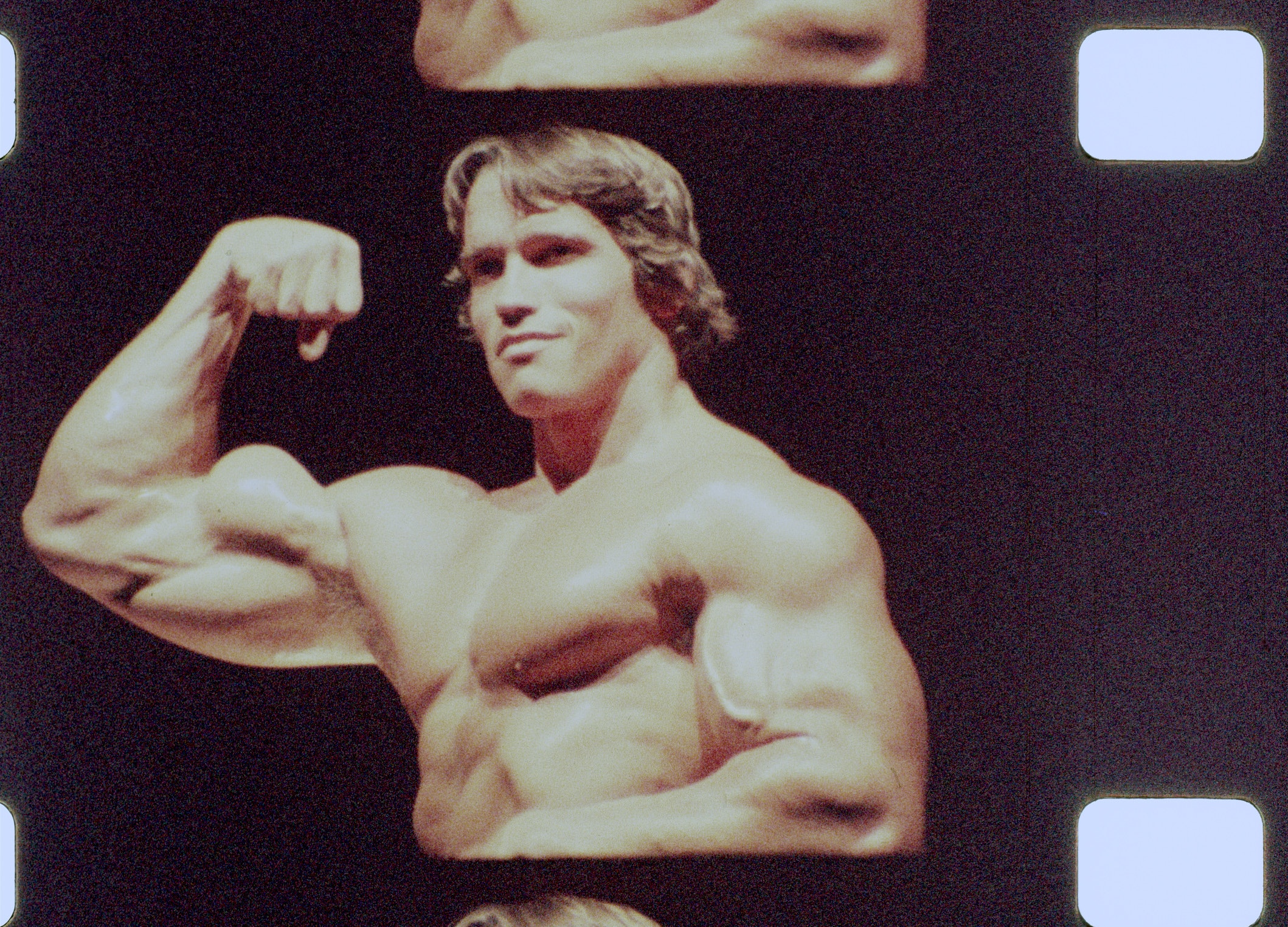 Film strip still of a young Arnold Schwarzenegger, shown from the waist up, shirtless and flexing his arm.
